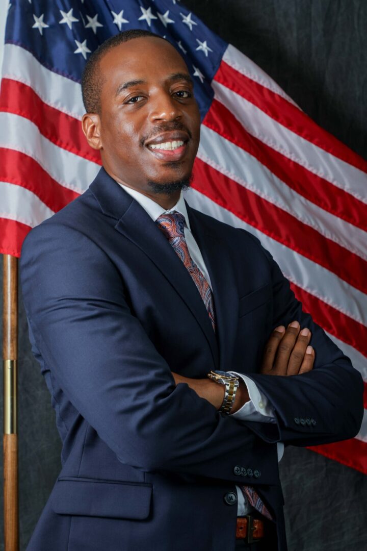 Confident man in a suit poses with crossed arms in front of an American flag.