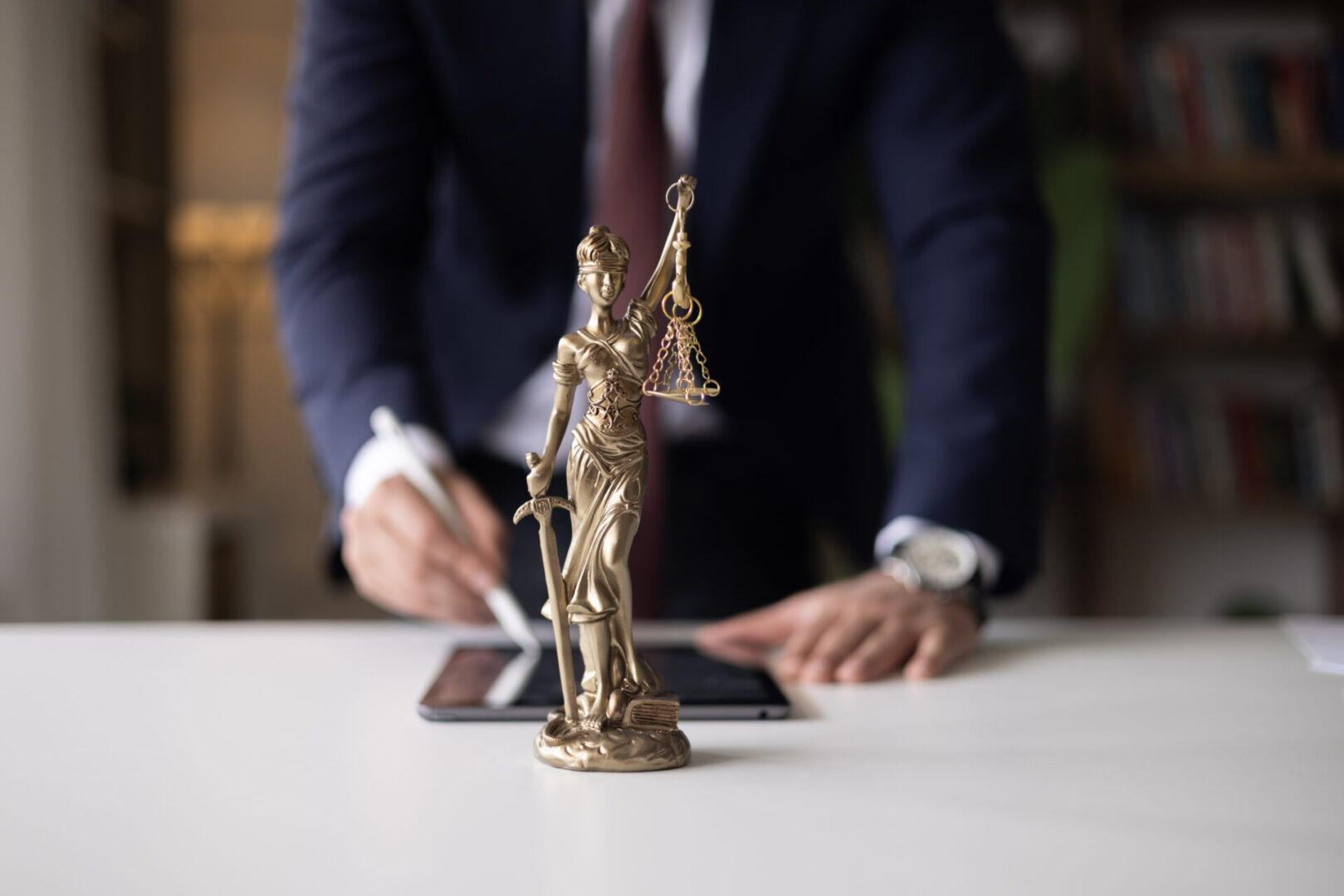 A bronze Lady Justice statue on a desk with a blurred lawyer in the background.